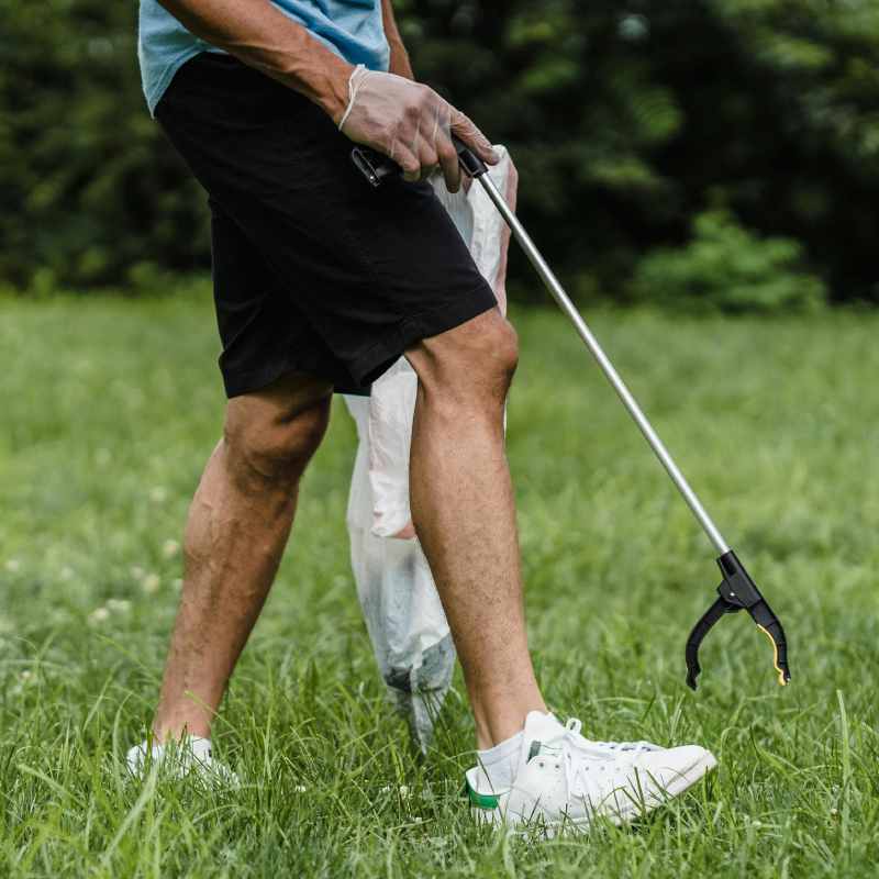 Lower half of a person walking in grass carrying a litter grabber and a plastic bag